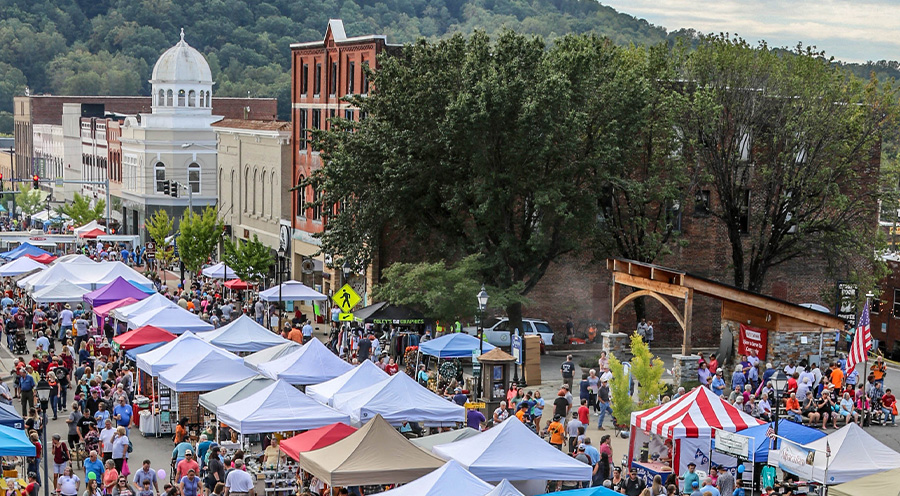 photo taken from a high elevation of a previous Mountain Glory festival with the historic Main Street filled with people browsing vendors, who are under teneted canopies. The bandstand can be seen to the right, and in the background the background is of forested hills